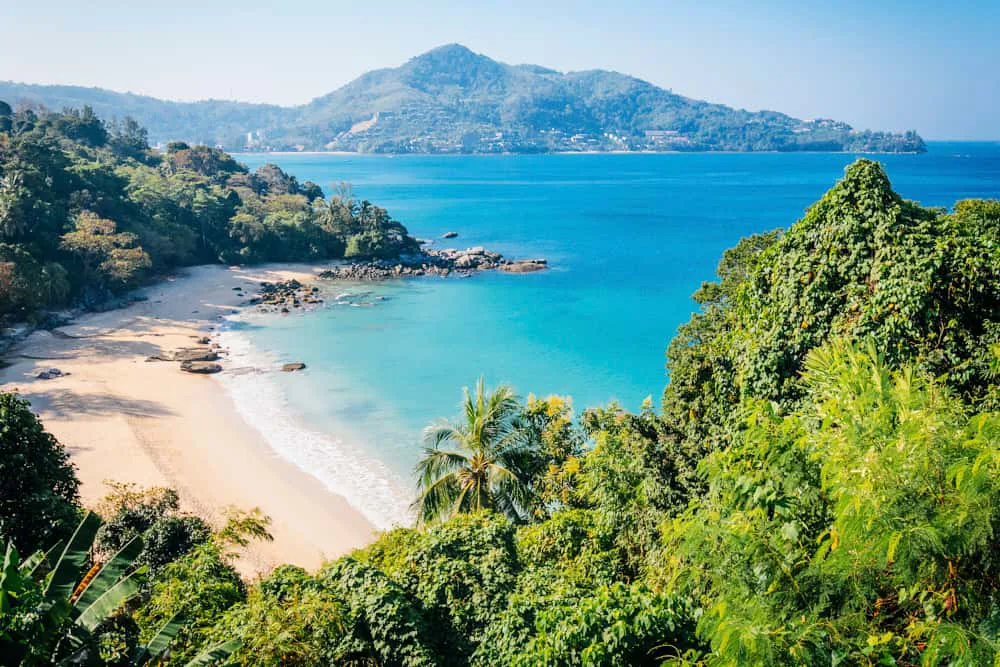 Panoramic view of Phuket's Patong Beach with turquoise waters, lush green hills, and colorful longtail boats at sunset.